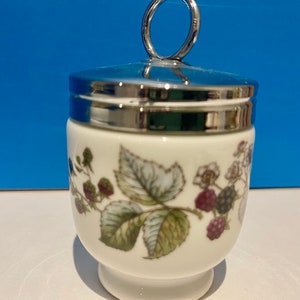 May include: White porcelain egg cup with a silver lid and a ring handle. The cup has a floral design with green leaves and purple berries.