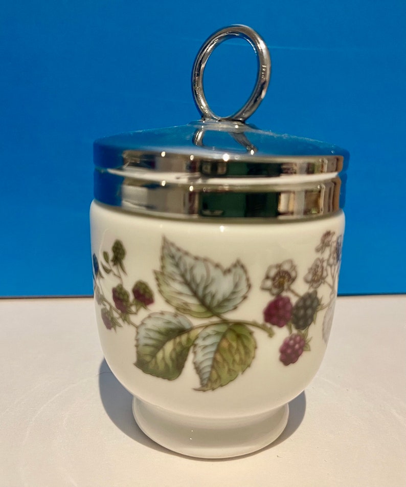 May include: White porcelain egg cup with a silver lid and a ring handle. The cup has a floral design with green leaves and purple berries.