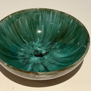 以下が含まれることがあります： 光沢のあるターコイズグリーンの釉薬を施したセラミックボウル。内側は、中央に向かって暗くなるテクスチャード加工された放射状のパターンが特徴です。縁は対照的な茶色のエッジです。この装飾的なボウルは、サービングまたはディスプレイに適しています。