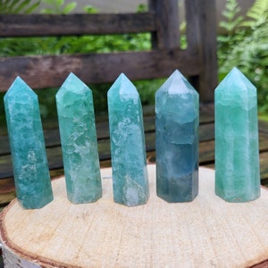 Op de afbeelding: Zes groene en blauwe fluorietkristallen op een houten oppervlak. De punten zijn allemaal verschillend van grootte en vorm, maar ze zijn allemaal gepolijst en hebben een gladde, glazige afwerking.