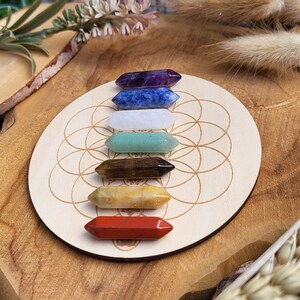 Könnte beinhalten: Ein Set aus sieben farbigen Kristallstäben, angeordnet auf einer Holzscheibe mit geometrischem Design. Die Kristalle sind in den Farbtönen Lila, Blau, Weiß, Grün, Braun, Gelb und Rot gehalten. Die Kristalle sind sechseckig.