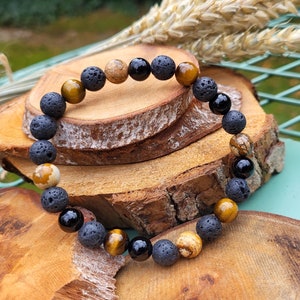 May include: A beaded bracelet featuring a mix of natural stone beads. The bracelet includes black lava rock beads, tiger's eye beads, black onyx beads, and jasper beads. The bracelet is displayed on a wooden surface.