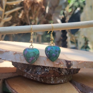 Peut inclure: Une paire de boucles d'oreilles pendantes avec des pendentifs en forme de cœur en pierre verte parsemée d'inclusions magenta. Les pendentifs sont suspendus à des crochets en métal doré, créant un design simple et élégant pour ces bijoux.