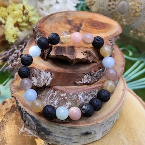 Könnte beinhalten: Ein mehrfarbiges Perlenarmband mit schwarzen Lavasteinen, hellblauen, rosa und gelben Perlen. Das Armband liegt auf einer Holzoberfläche.