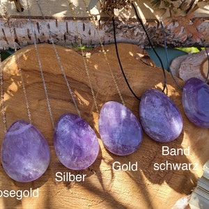 Puede incluir: Cinco colgantes de piedra de amatista en cadenas, cada uno con una cadena de color diferente: oro rosa, plata, oro, negro y gris. Las piedras son todas de la misma forma y tamaño, y todas están colocadas sobre un fondo de madera.