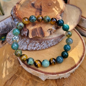 Op de afbeelding: Een groene, bruine en gouden kralen armband met een zilveren bedel. De armband is gemaakt van natuurlijke stenen en ligt op een houten oppervlak.