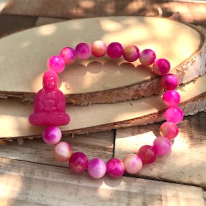 May include: A pink and white agate beaded bracelet with a pink Buddha charm. The bracelet is on a wooden surface.