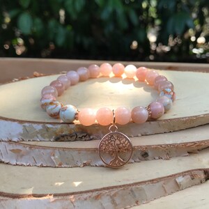Puede incluir: Una pulsera de cuentas con piedras lisas y redondas de color melocotón y varias cuentas jaspeadas en azul y blanco. La pulsera incluye un dije circular de tono oro rosa que representa un árbol con ramas y raíces detalladas, suspendido de una pequeña argolla.