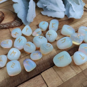 Könnte beinhalten: Ein Set aus 20 weißen Opal-Steinen mit goldenen Runen, die auf jedem Stein eingraviert sind. Die Steine sind auf einer Holzoberfläche angeordnet.
