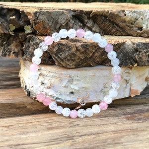 May include: A beaded bracelet featuring alternating pink and white round beads. The bracelet is made with a stretch cord and has a small silver-colored charm. The beads have a smooth, polished finish.