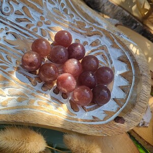 Könnte beinhalten: Eine Sammlung von 12 glatten, runden, rosa und violetten Edelsteinkugeln. Die Kugeln sind in einem Cluster auf einem Holztablett mit einem weißen Blumenmuster angeordnet.