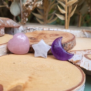 Könnte beinhalten: Drei kleine Kristallsteine, die in die Formen eines Herzens, eines Sterns und eines Halbmondes geschnitzt sind. Das Herz ist rosa, der Stern ist weiß und der Mond ist lila. Die Steine liegen auf einer Holzoberfläche.