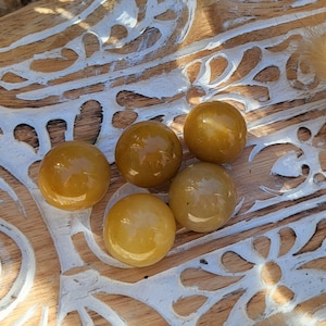 Może przedstawiać: Pięć gładkich, okrągłych, polerowanych kamieni w kolorze złotożółtym. Kamienie są ułożone w grupie na drewnianej powierzchni z białym wzorem kwiatowym.