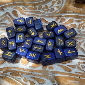 Peut inclure: Ensemble de pierres de runes bleu foncé avec des symboles dorés. Les pierres sont dispersées sur une surface en bois sculpté, créant une esthétique mystique. Les runes sont probablement utilisées pour la divination ou les pratiques spirituelles.