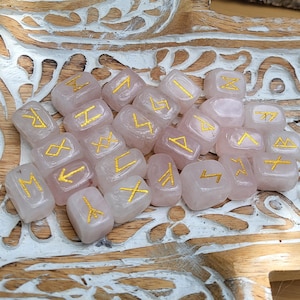 Peut inclure: Un ensemble de runes en quartz rose avec des symboles dorés. Les runes sont disposées sur un plateau en bois sculpté avec un motif peint en blanc. Les runes sont de couleur rose clair et les symboles sont dorés.