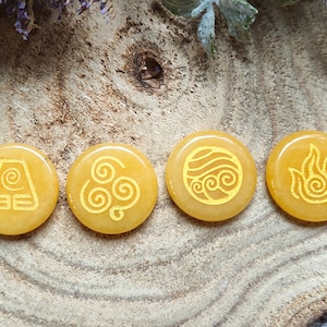 Könnte beinhalten: Vier gelbe runde Steine mit verschiedenen Symbolen, die in Gold eingeätzt sind. Die Symbole sind die vier Elemente aus Avatar – Der Herr der Elemente: Erde, Luft, Wasser und Feuer.