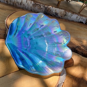 May include: Iridescent blue seashell-shaped dish. The dish has a scalloped edge and a textured surface, reflecting a spectrum of colors. The dish is made of glass and is a decorative item.