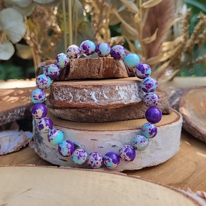 May include: A bracelet made of round beads in shades of purple, teal, and cream. The beads are displayed on a wooden surface. The bracelet is a piece of jewellery. The beads are approximately 1 cm in diameter.