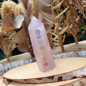 Op de afbeelding: Een heldere roze rozenkwarts kristallen punt met de zeven chakra symbolen erop gegraveerd. De symbolen zijn in regenboogkleuren. Het kristal ligt op een houten oppervlak.