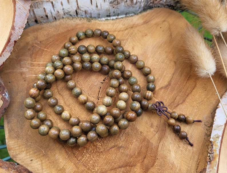 May include: A wooden mala bead necklace with brown and green beads. The necklace is made of natural wood and has a knotted tassel.