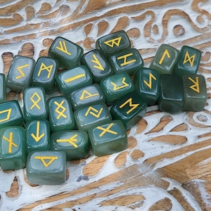 May include: A collection of green, translucent rune stones with gold symbols. The stones are scattered on a carved wooden surface, creating a mystical aesthetic. The runes are used for divination and spiritual practices.