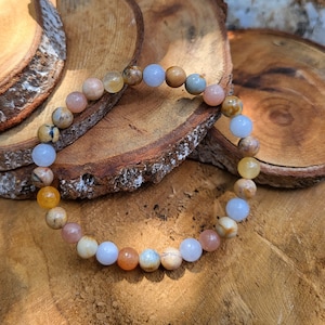 Puede incluir: Un brazalete de cuentas multicolores con cuentas marrones, naranjas, rosas, azules y blancas. El brazalete está hecho de piedras naturales y está sobre una superficie de madera.