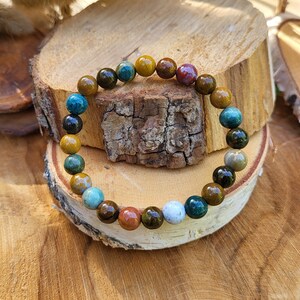 May include: A multi-coloured gemstone bracelet. The bracelet is made up of polished beads in shades of blue, green, brown, yellow, and red. The bracelet is displayed on a wooden surface, highlighting the natural colours of the stones.