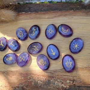 May include: Twelve purple amethyst stones with gold symbols on a wooden surface. The symbols include a sun, moon, eye, cross, and other abstract designs.
