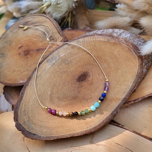 Op de afbeelding: Een delicate ketting met een reeks kleurrijke edelsteen kralen. De kralen bevatten tinten roze, groen, blauw en paars, met goudkleurige accenten. De ketting wordt gepresenteerd op een rustiek houten oppervlak.