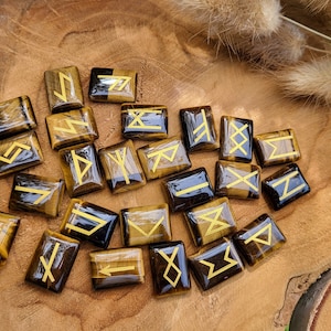 Peut inclure: Un ensemble de 20 pierres de runes en œil de tigre avec des runes dorées sur chaque pierre. Les pierres sont disposées sur une surface en bois.