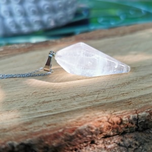 May include: A clear quartz crystal pendant on a silver chain. The crystal is faceted and elongated, with a pointed end. The chain is delicate, and the pendant is displayed on a wooden surface.