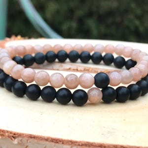 May include: Two beaded bracelets. One bracelet features peach-colored beads, and the other has black beads. The bracelets are stacked on top of each other. The background is a blurred green and wood surface.