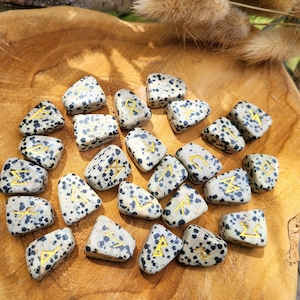 Peut inclure: Un ensemble de 20 pierres de jaspe dalmatien avec des runes dorées peintes dessus. Les pierres sont disposées sur une surface en bois.