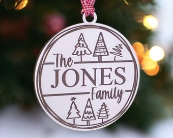 May include: A round, silver-colored Christmas ornament with the text "The JONES Family" and tree illustrations. The ornament hangs from a red and white checkered ribbon, displayed on a Christmas tree.