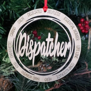 May include: A wooden Christmas ornament with the word "Dispatcher" cut out in the center. The ornament has a circular design with words like "Caring", "Calm", "Dedicated", and "Essential Worker" engraved around the edge. It hangs from a red ribbon.