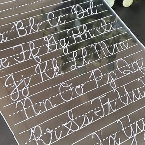 May include: A clear acrylic cursive handwriting practice board with white dotted lines and letters from A to Z. The letters are in a cursive style, with both uppercase and lowercase letters displayed. The board is angled, with a dark background.
