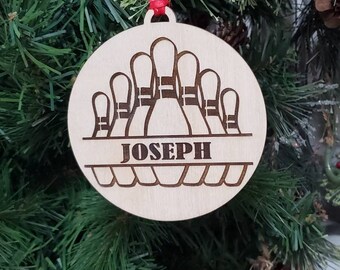May include: A round, wooden Christmas ornament featuring a bowling pin design. The ornament has the name "JOSEPH" engraved in a horizontal band. A red ribbon is attached for hanging. The ornament is displayed on a Christmas tree.