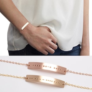 May include: Two gold bracelets with a delicate chain and a curved bar charm. The charms are engraved with Morse code.
