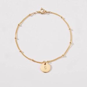 Może przedstawiać: Złota bransoletka z łańcuszkiem i małym okrągłym wisiorkiem z wygrawerowaną literą "S".