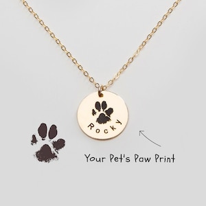 May include: Gold necklace with a round pendant featuring a paw print and the name "Rocky" engraved on it. The pendant is hanging from a delicate gold chain.