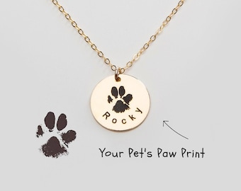 May include: Gold necklace with a round pendant featuring a paw print and the name "Rocky" engraved on it. The pendant is hanging from a delicate gold chain.