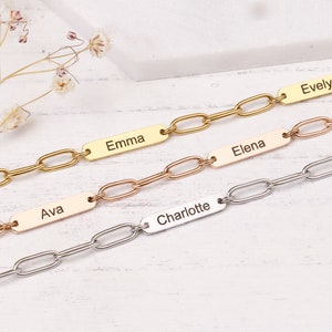May include: Four personalized chain link bracelets in gold, rose gold, and silver. Each bracelet features a rectangular nameplate with a different name engraved: Liam, Emma, Ava, Elena, Evelyn, and Charlotte. The bracelets are displayed on a light-colored surface.