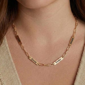May include: A gold-colored chain necklace with rectangular nameplates. Each plate has a name engraved in black. The names visible are Liam, Emma, Evelyn, and Amelia. The necklace is worn around the neck.