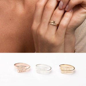 May include: Gold-coloured wrap ring with engraved text. The ring is shown on a person's finger, and additional rings in rose gold and silver are displayed below. The gold ring has the initials "M S" and Roman numerals. The rose gold ring is engraved with "Sophia" and "Ethan".