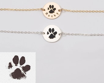 May include: Two silver necklaces with a round pendant featuring a black paw print. The top necklace has the word "Rocky" engraved on the pendant.