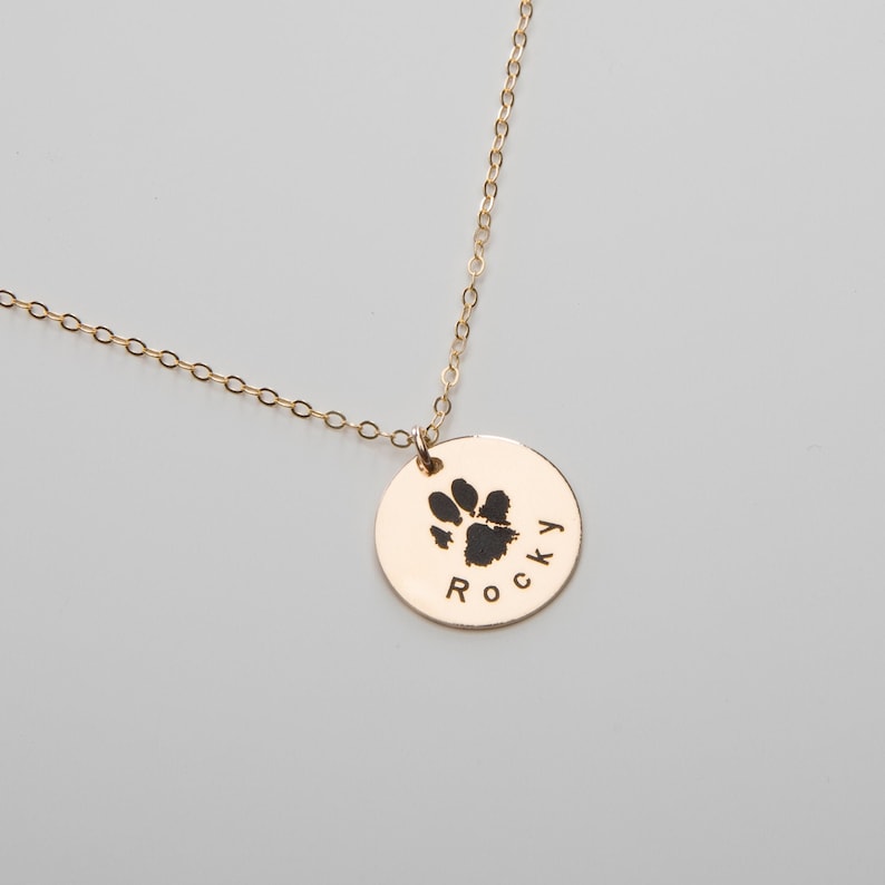 May include: Gold necklace with a round pendant featuring a black paw print and the name "Rocky" engraved on it.