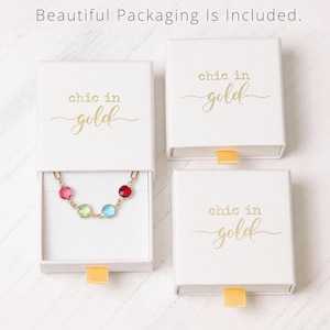 May include: Three white square jewellery boxes with gold text reading chic in gold. One box is open, revealing a gold chain bracelet featuring four round faceted stones in red, light green, light blue, and pink, each set in a gold-toned metal frame.