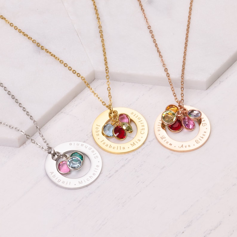 May include: Three gold necklaces with personalized engraved circles and colorful crystal charms. The necklaces are on a white marble background. The circles are engraved with names, including "John, Isabella, Mia, Charlotte", "Liam, Ava, Ella, Olivia", and "Abigail, Michelle, Sydney".