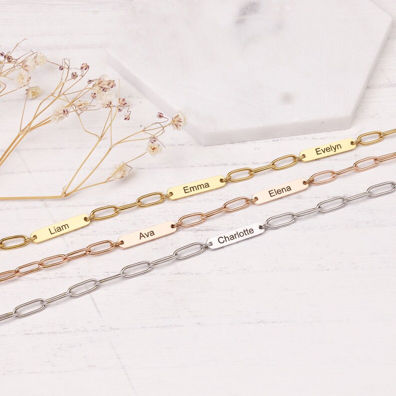 May include: Four personalized chain link bracelets in gold, rose gold, and silver. Each bracelet features a rectangular nameplate with a different name engraved: Liam, Emma, Ava, Elena, Evelyn, and Charlotte. The bracelets are displayed on a light-colored surface.