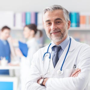 May include: A smiling doctor with gray hair and a beard, wearing a white coat, blue tie, and stethoscope. The doctor has their arms crossed. Other people are blurred in the background.